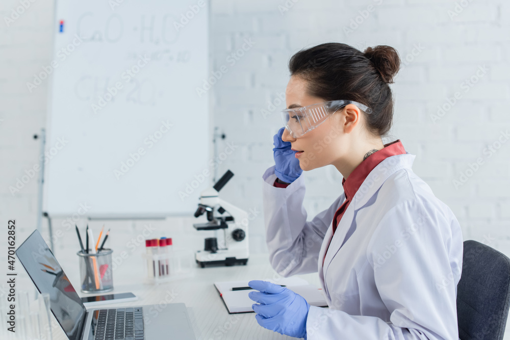 side view of tattooed scientist in white coat and goggles looking at laptop near test tubes. side view of tattooed scientist in white coat and goggles looking at laptop near test tubes.