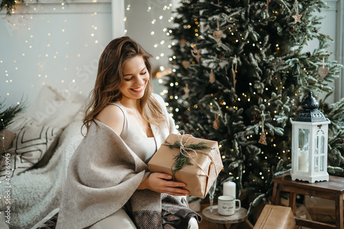 Smiling sincere beautiful young woman in comfortable light winter clothes holds out a wrapped gift box to the camera, prepares a Christmas present, celebrates the winter holidays