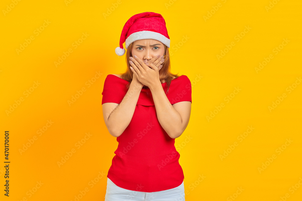 Surprised young Asian woman wearing Christmas hat covering mouth with hands on yellow background Surprised young Asian woman wearing Christmas hat covering mouth with hands on yellow background