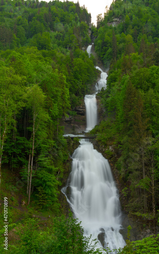 Giant waterfall falling between abundant green vegetation and in the center a suspension bridge · Giessbach (Austria) · Vertical view