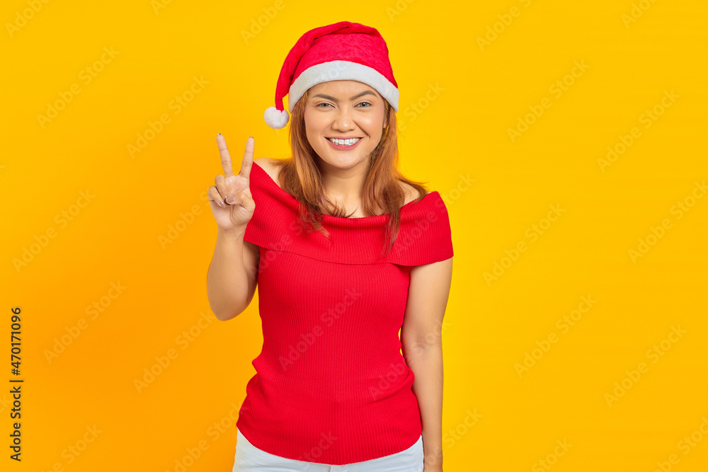 Cheerful young Asian woman wearing Christmas hat showing peace sign on yellow background Cheerful young Asian woman wearing Christmas hat showing peace sign on yellow background