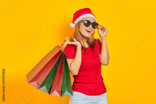Smiling young Asian woman in Santa Claus hat and sunglasses holding shopping bags isolated over yellow background