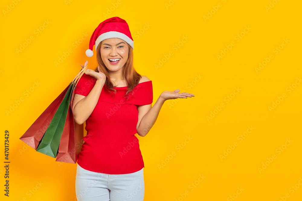 Cheerful young Asian woman in Santa Claus hat holding shopping bags and showing copy space in palms on yellow background Cheerful young Asian woman in Santa Claus hat holding shopping bags and showing copy space in palms on yellow background