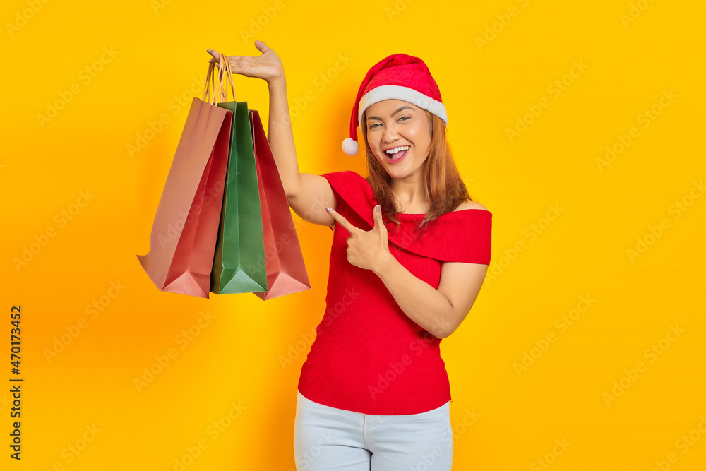 Cheerful young Asian woman in Santa Claus hat pointing finger at shopping bag on yellow background Cheerful young Asian woman in Santa Claus hat pointing finger at shopping bag on yellow background