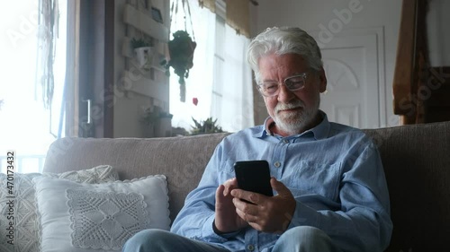 One old man using phone at home sitting on the sofa smiling and having fun learning chatting with friends and surfing on the web. Satisfied male senior indoor with smartphone.
