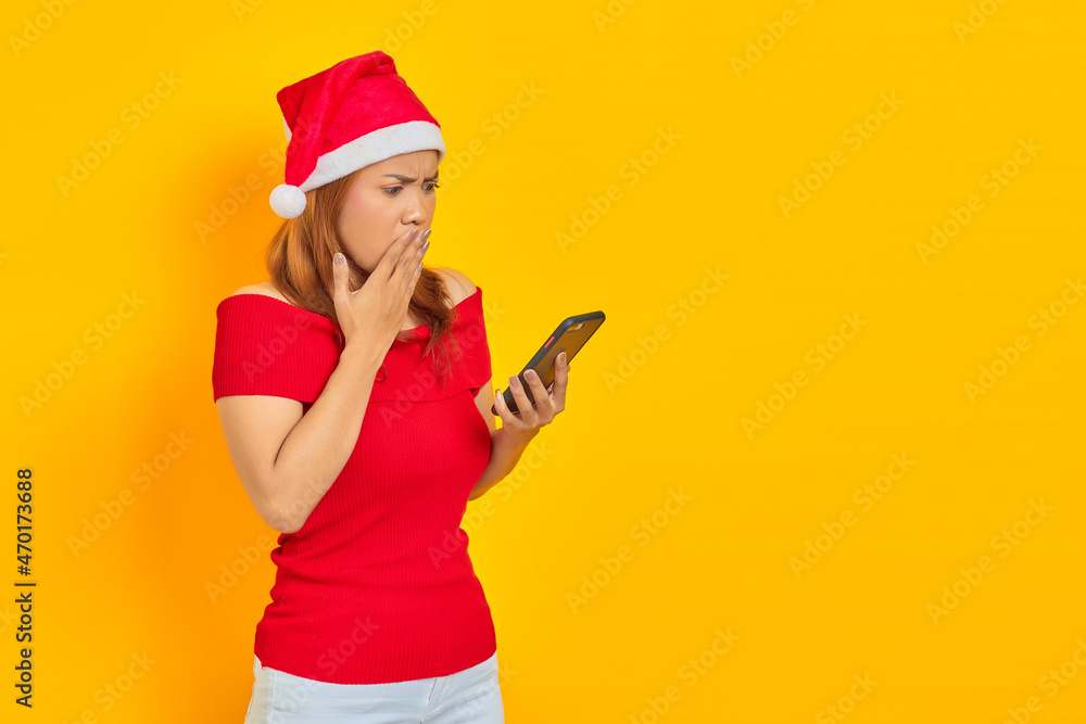Surprised young Asian woman in Santa Claus hat looking at mobile phone with open mouth isolated over yellow background Surprised young Asian woman in Santa Claus hat looking at mobile phone with open mouth isolated over yellow background