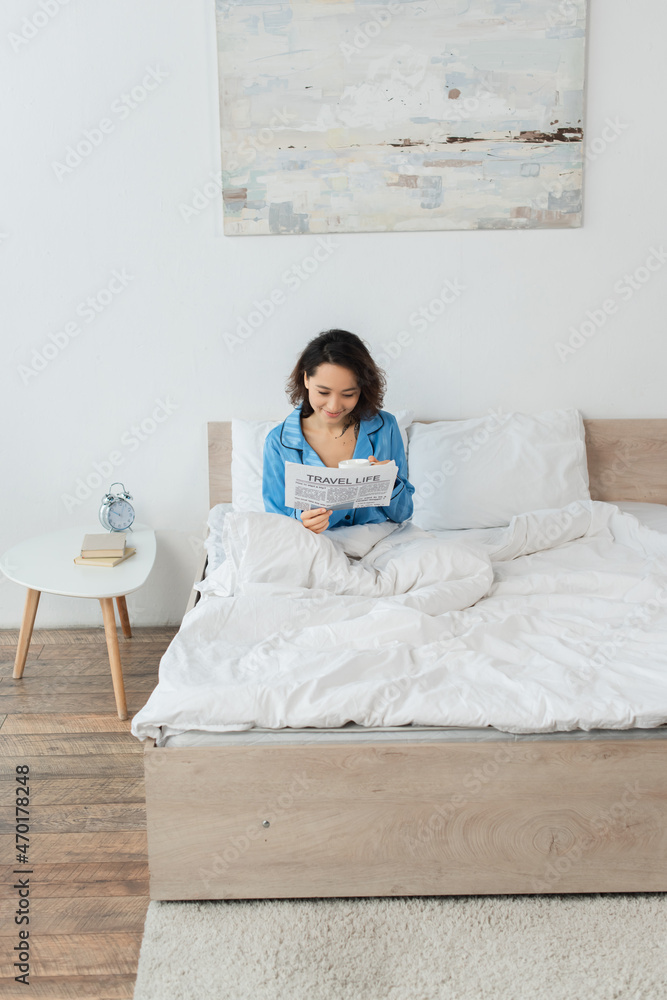 cheerful young woman in pajamas reading travel life newspaper and holding cup in bed.
