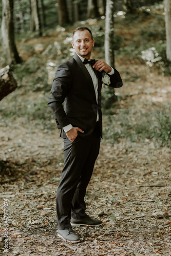 The groom in a black suit poses for a photo in the green garden