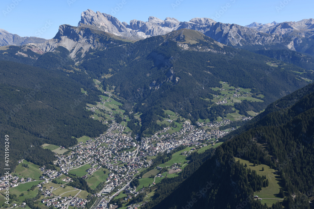 Luftaufnahme vom Ort Sankt St. Ulrich Gröden im Grödnertal. Die Amtssprachen sind Deutsch, Italienisch und Ladinisch.  Im Hintergrund das Bergmassiv der Geislerspitzen.  