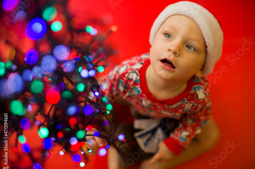 baby in santa claus hat on a red background looks at the colorful lights from garlands.