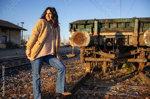 Portrait of a beautiful young woman in happy dawn on the dangerous railroad tracks. Woman in boots and jeans on the train tracks. Beautiful woman enjoying the train tracks beside a train carriage.