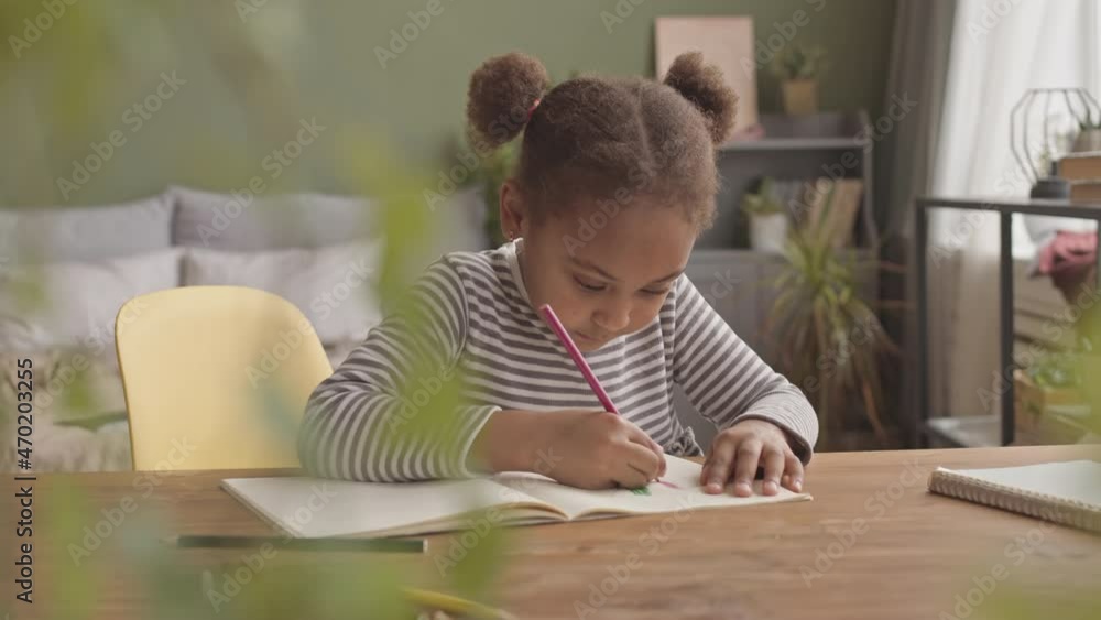 Waist up slowmo shot of little African-American girl drawing in copybook sitting at desk in cozy bedroom Waist up slowmo shot of little African-American girl drawing in copybook sitting at desk in cozy bedroom