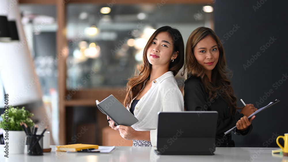 Two attractive female colleagues sitting in office and smiling to camera. Two attractive female colleagues sitting in office and smiling to camera.