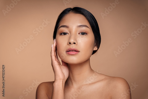 Fotografie Closeup headshot portrait of young nude asian woman touching smooth clean fresh cheek skin looking at camera