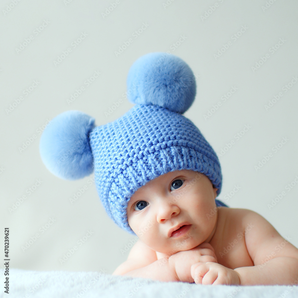 Sweet newborn baby in a blue hat with pompoms looks into the camera. A calm two-month-old infant is lying on a blanket. A beautiful, cute little child with blue eyes looks into the camera. . Sweet newborn baby in a blue hat with pompoms looks into the camera. A calm two-month-old infant is lying on a blanket. A beautiful, cute little child with blue eyes looks into the camera. .