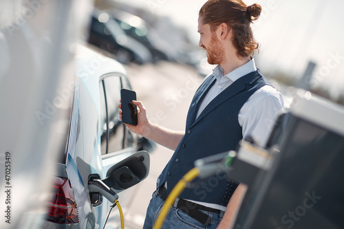 Smiling man is holding a phone while electric car charged at the charging station