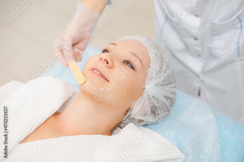 Hands of cosmetology specialist applying facial mask using stick  making skin hydrated and face glowing. Attractive woman relaxing smiling and enjoying spa procedures