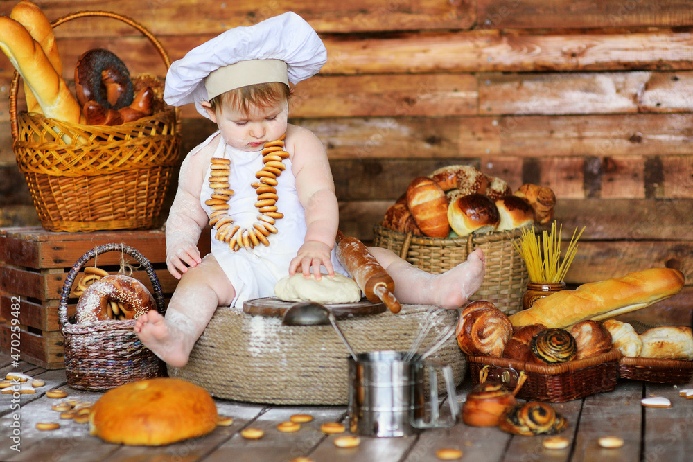a baby baker boy in a chef's hat and apron smeared in flour with a bunch of bagels around his neck prepares dough for baking.