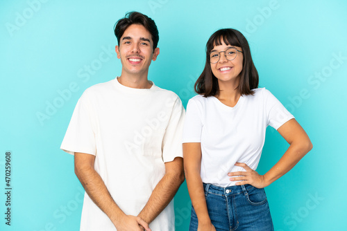 Young mixed race couple isolated on blue background posing with arms at hip and smiling
