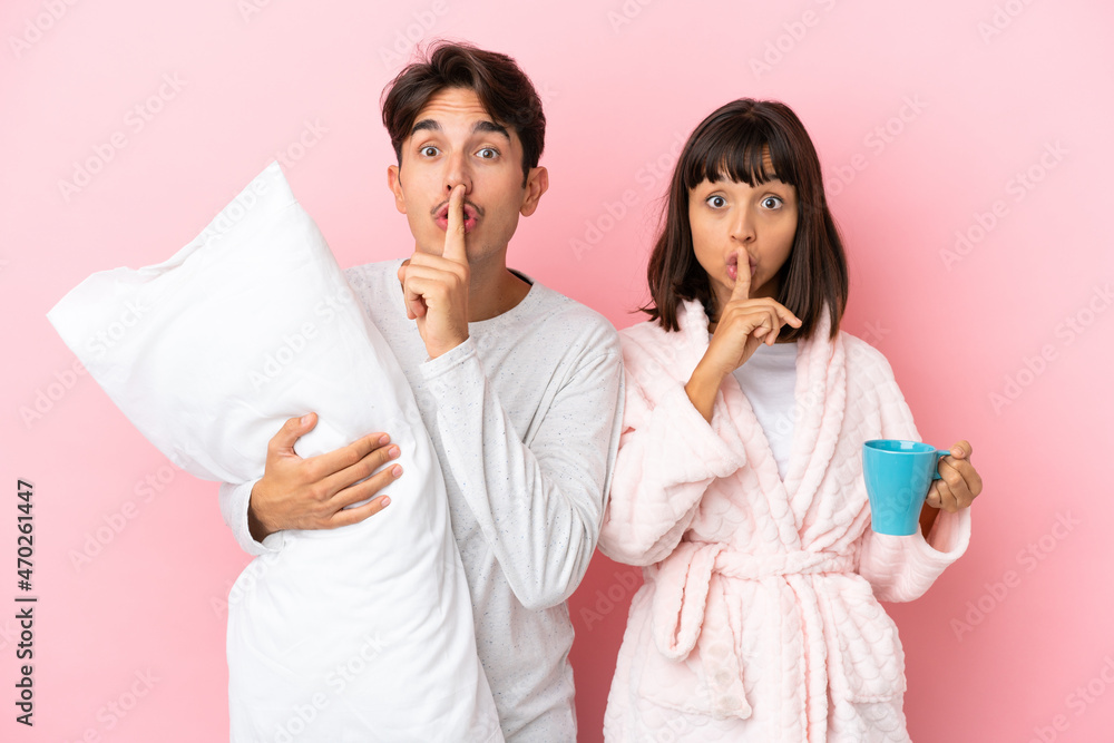 Young couple in pajamas isolated on pink background showing a sign of silence gesture putting finger in mouth