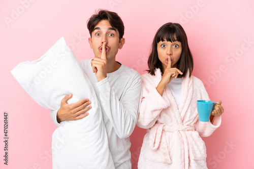 Young couple in pajamas isolated on pink background showing a sign of silence gesture putting finger in mouth