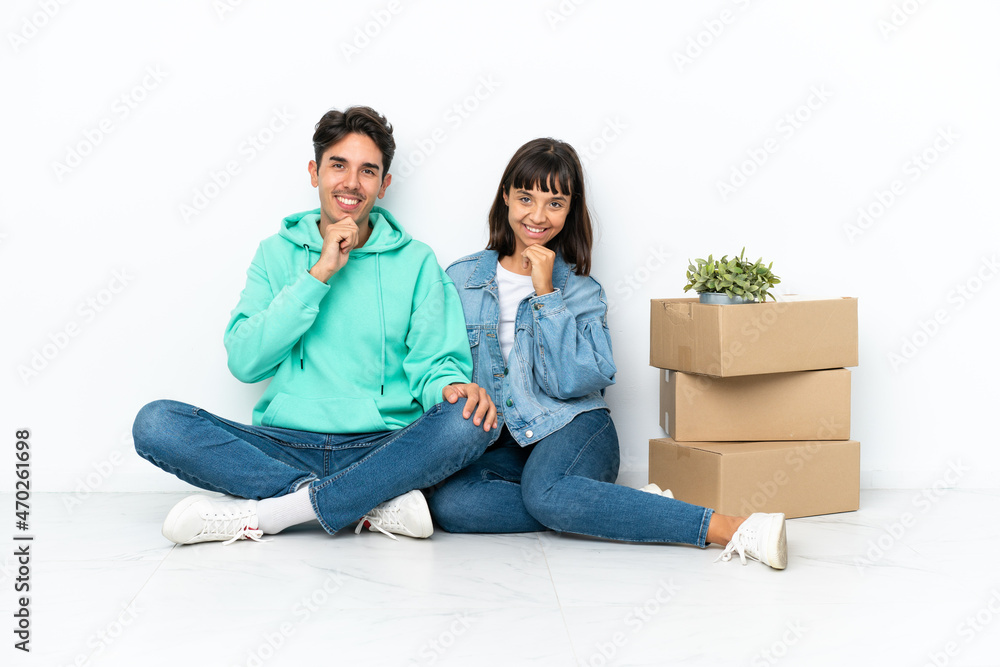 Young couple making a move while picking up a box full of things sitting on the floor isolated on white background smiling and looking to the front with confident face Young couple making a move while picking up a box full of things sitting on the floor isolated on white background smiling and looking to the front with confident face