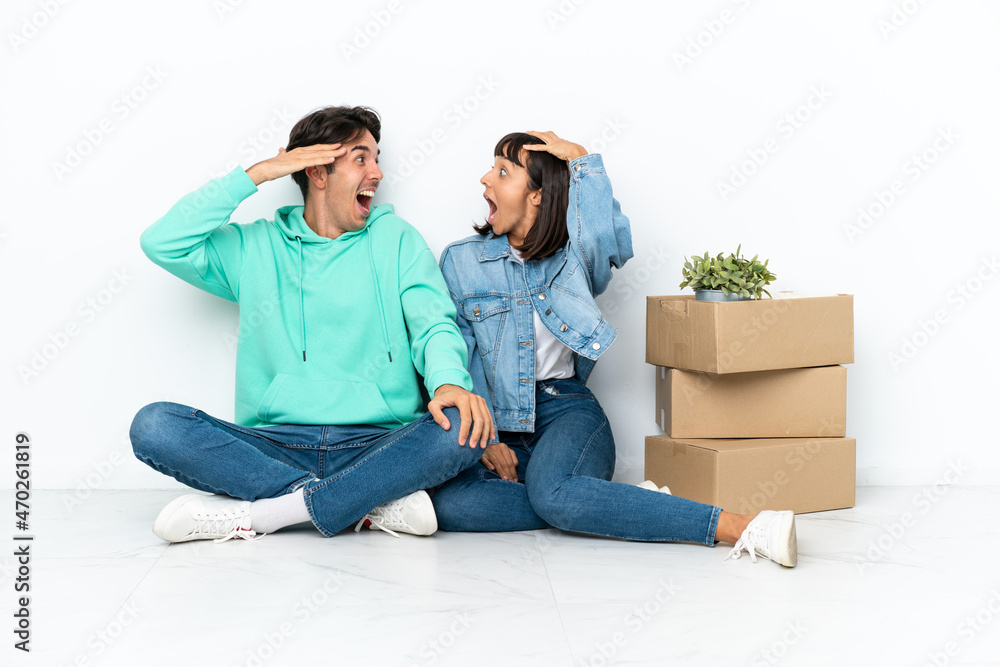 Young couple making a move while picking up a box full of things sitting on the floor isolated on white background with surprise and shocked facial expression Young couple making a move while picking up a box full of things sitting on the floor isolated on white background with surprise and shocked facial expression