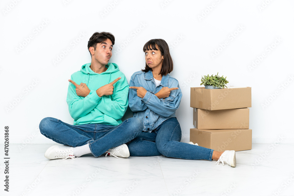 Young couple making a move while picking up a box full of things sitting on the floor isolated on white background pointing to the laterals having doubts Young couple making a move while picking up a box full of things sitting on the floor isolated on white background pointing to the laterals having doubts