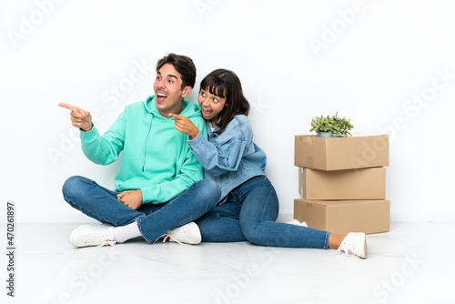 Young couple making a move while picking up a box full of things sitting on the floor isolated on white background presenting an idea while looking smiling towards © luismolinero