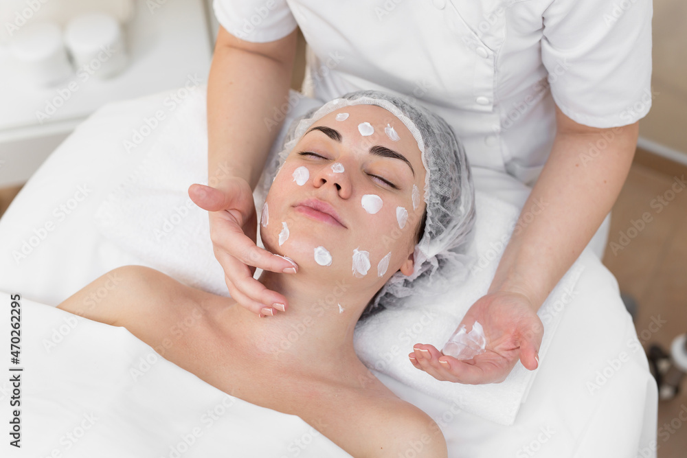 Naklejka premium Hands of beautician apply cream to face of woman. Concept of beauty and youth. Close-up beautician causes moisturizer on the girl's face