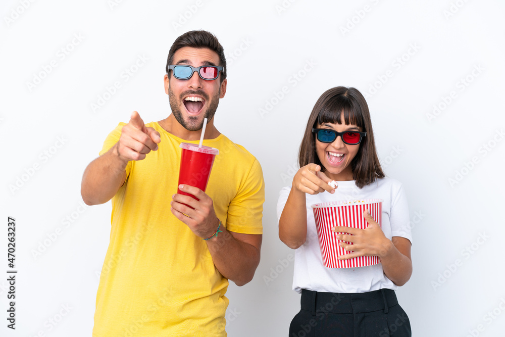 Young couple isolated on white background with 3d glasses and holding a big bucket of popcorns while pointing away Young couple isolated on white background with 3d glasses and holding a big bucket of popcorns while pointing away