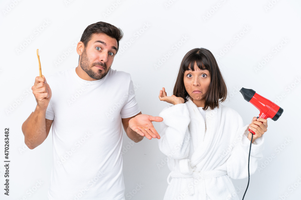Young caucasian couple holding a hairdryer and toothbrush isolated on white background having doubts while raising hands and shoulders Young caucasian couple holding a hairdryer and toothbrush isolated on white background having doubts while raising hands and shoulders
