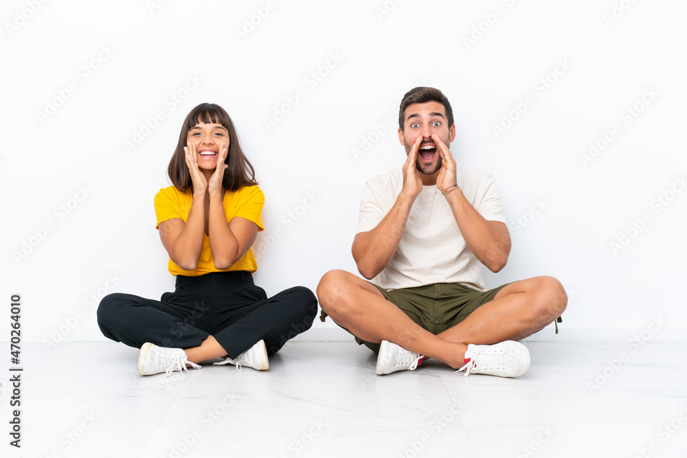Young couple sitting on the floor isolated on white background shouting and announcing something Young couple sitting on the floor isolated on white background shouting and announcing something