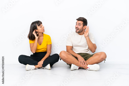 Young couple sitting on the floor isolated on white background listening to something by putting hand on the ear