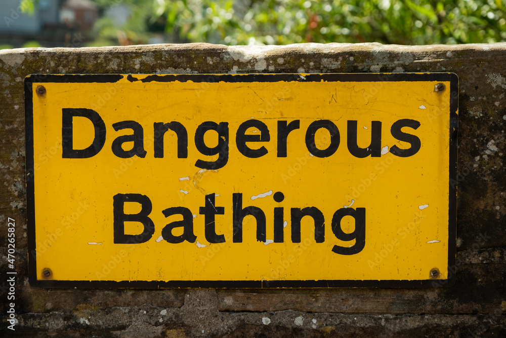 A yellow dangerous bathing sign fixed to a stone wall beside water A yellow dangerous bathing sign fixed to a stone wall beside water
