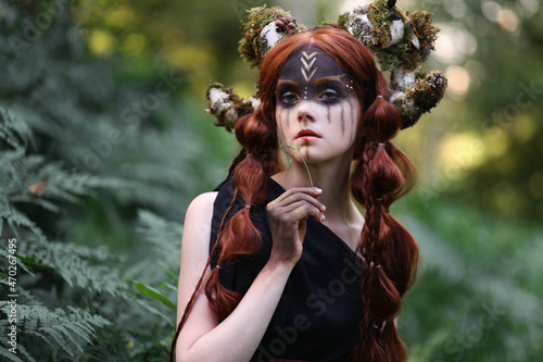 Beautiful red haired girl with braided hair and dark faceart with mossy horns among fern. Faitytale portrait of faun in dark forest photo