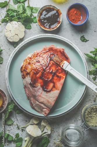 Raw meat with homemade marinade on blue plate with garlic, spices and herbs on grey concrete background. Meat preparation for BBQ or grill. Top view.