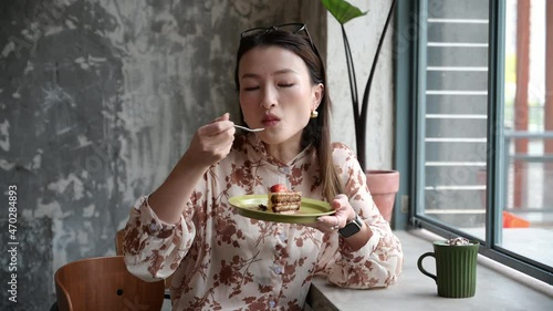 Beautiful Chinese Asian young woman visits cafe to have her dessert. Portrait of stunning girl eating piece of cake and enjoying mocha latte coffee. Business lady having coffee break in cafeteria.