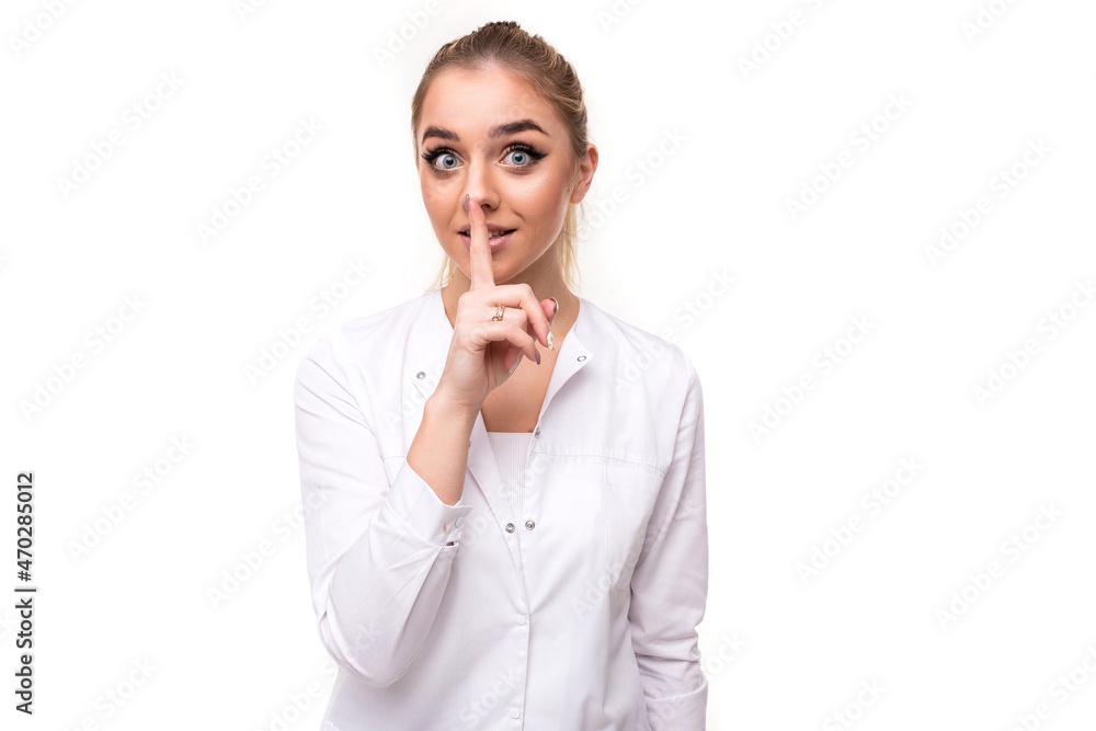Photo of a beautiful pretty cute attractive positive young blonde woman with a ponytail and makeup in a white medical coat of a nurse showing shhh and secret gesture with one finger isolated on a Photo of a beautiful pretty cute attractive positive young blonde woman with a ponytail and makeup in a white medical coat of a nurse showing shhh and secret gesture with one finger isolated on a