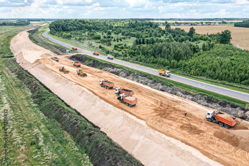 panoramic aerial view of large road construction site with working heavy machines - bulldozer, grader, earthmover and dump trucks