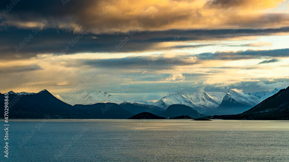 Wilde, schneebedeckte Berge in der Umgebung von Ørnes, Norwegen, im orangen Morgenlicht bei Sturm mit Wolken. Schneefahnen wehen von den Bergspitzen herunter. Kreuzfahrt mit dem Postschiff 