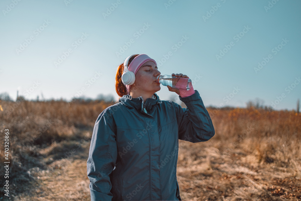 Active young woman in sportswear and wireless headphones drinks fresh water from a bottle in natural autumn park banner background Active young woman in sportswear and wireless headphones drinks fresh water from a bottle in natural autumn park banner background