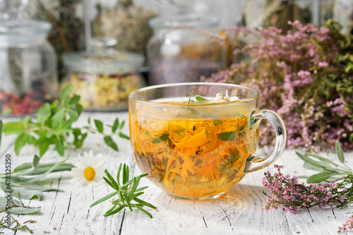 Glass cup of healthy calendula herbal tea, heather, rosemary, chamomile and salvia medicinal herbs and flowers on table. Jars of dried medicinal herbs on background. Alternative herbal medicine.