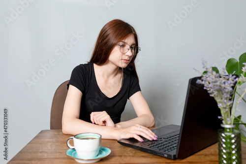 young woman is sitting in front of laptop, shopping on Internet, chatting with friends or just surfing web. cup of coffee on table