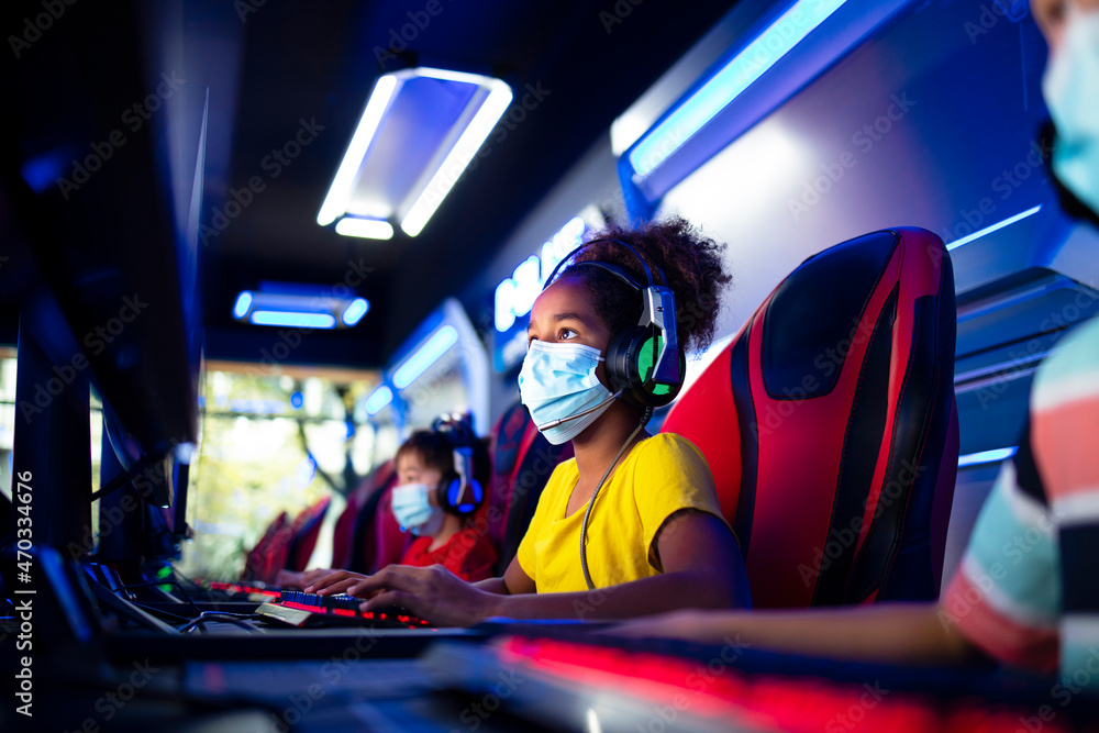 Obraz premium Portrait of an African American girl wearing face mask and headset playing video games on computer in game room during corona virus pandemic.