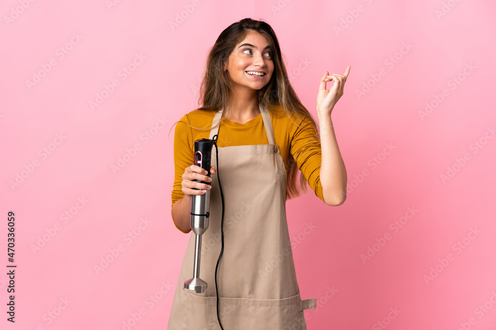 Young chef woman using hand blender isolated on pink background pointing up a great idea Young chef woman using hand blender isolated on pink background pointing up a great idea