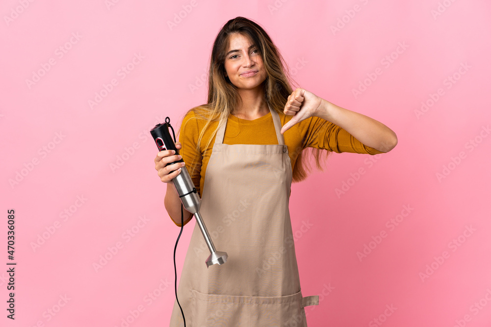 Young chef woman using hand blender isolated on pink background showing thumb down with negative expression