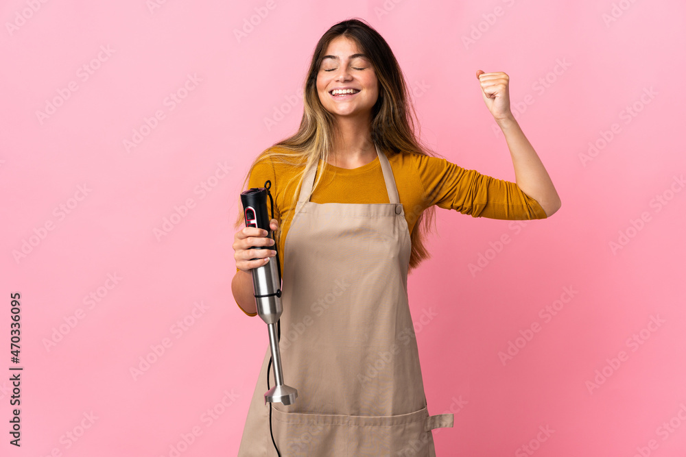 Young chef woman using hand blender isolated on pink background doing strong gesture Young chef woman using hand blender isolated on pink background doing strong gesture