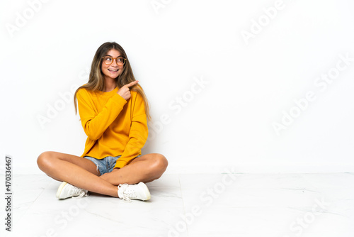 Young woman sitting on the floor isolated on white background pointing to the side to present a product