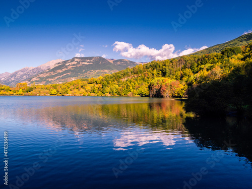 Lago de montaña con bosque en otoño y cielo azul con nubes blancas reflejadas en el agua.
